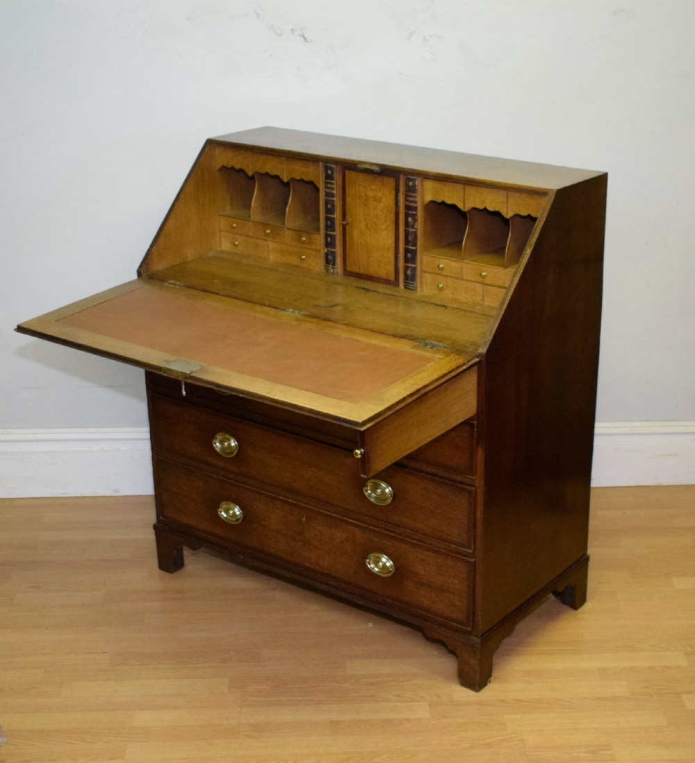 A Georgian oak bureau featuring a slanted fall-front lid above four graduated drawers fitted with brass oval handles and raised on bracket feet. View XXVI