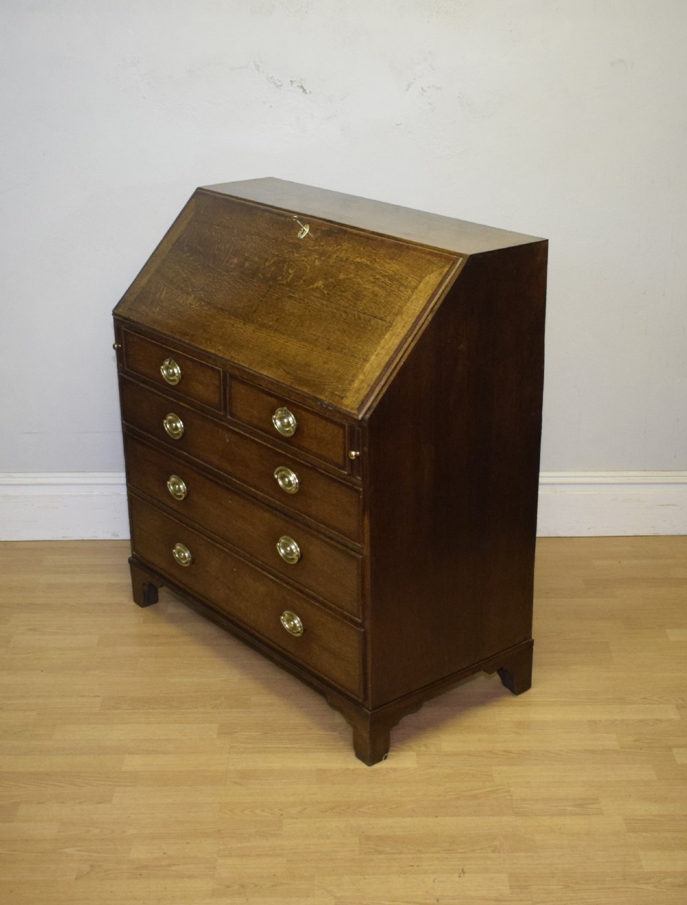 A Georgian oak bureau featuring a slanted fall-front lid above four graduated drawers fitted with brass oval handles and raised on bracket feet. View XXVII