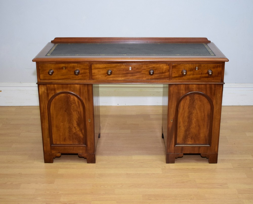 A Victorian mahogany pedestal desk featuring a dark green leather inset writing surface, three frieze drawers, and arched panel pedestal doors.