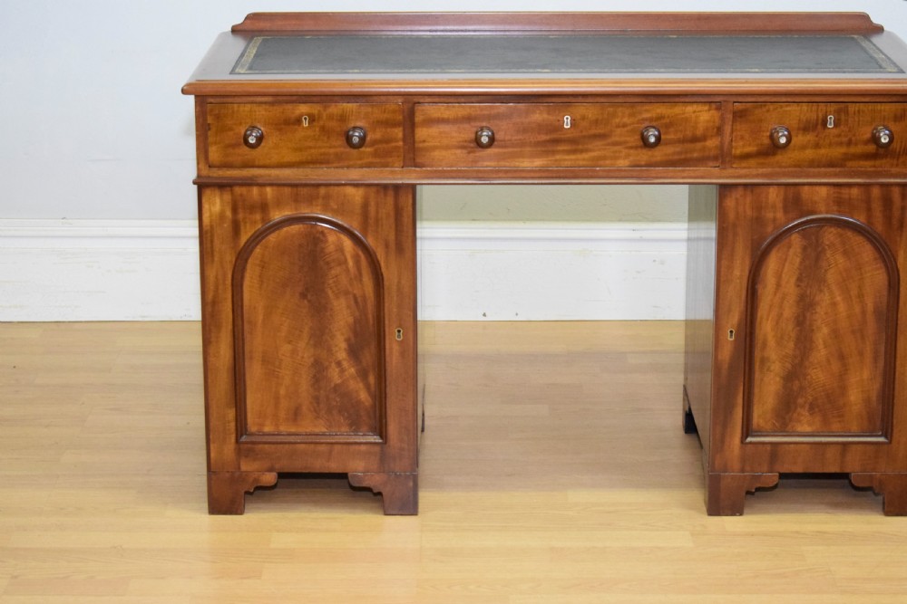 A Victorian mahogany pedestal desk featuring a dark green leather inset writing surface, three frieze drawers, and arched panel pedestal doors. View V