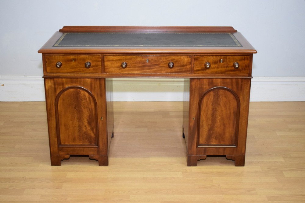 A Victorian mahogany pedestal desk featuring a dark green leather inset writing surface, three frieze drawers, and arched panel pedestal doors. View VI