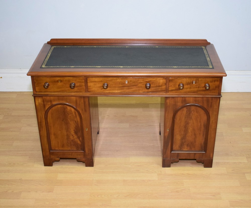 A Victorian mahogany pedestal desk featuring a dark green leather inset writing surface, three frieze drawers, and arched panel pedestal doors. View VII