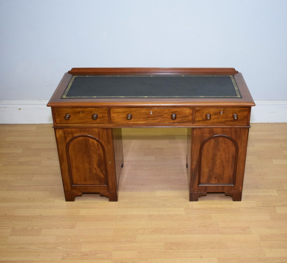 A Victorian mahogany pedestal desk featuring a dark green leather inset writing surface, three frieze drawers, and arched panel pedestal doors. View VIII