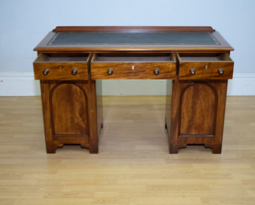 A Victorian mahogany pedestal desk featuring a dark green leather inset writing surface, three frieze drawers, and arched panel pedestal doors. View X