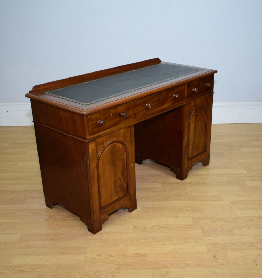 A Victorian mahogany pedestal desk featuring a dark green leather inset writing surface, three frieze drawers, and arched panel pedestal doors. View XIII