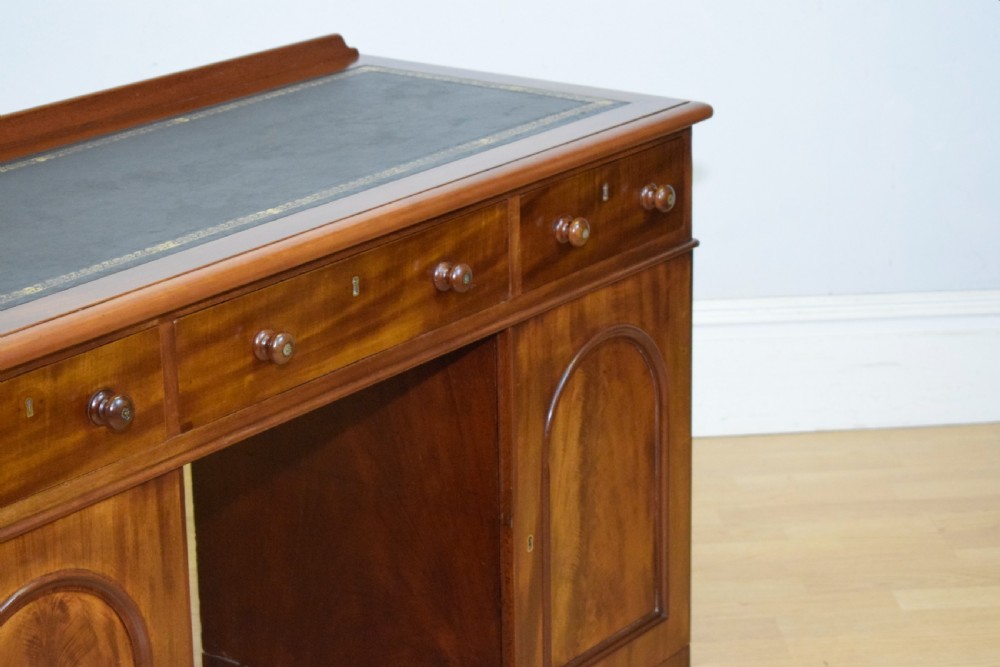 A Victorian mahogany pedestal desk featuring a dark green leather inset writing surface, three frieze drawers, and arched panel pedestal doors. View XIV