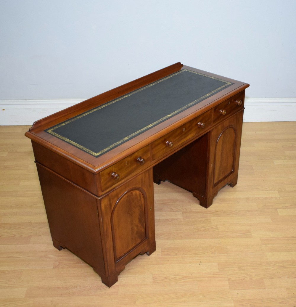 A Victorian mahogany pedestal desk featuring a dark green leather inset writing surface, three frieze drawers, and arched panel pedestal doors. View XV