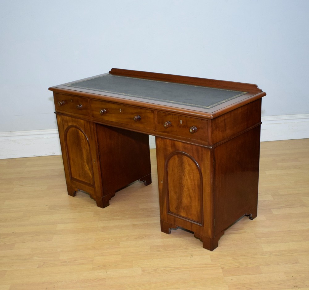 A Victorian mahogany pedestal desk featuring a dark green leather inset writing surface, three frieze drawers, and arched panel pedestal doors. View XVI
