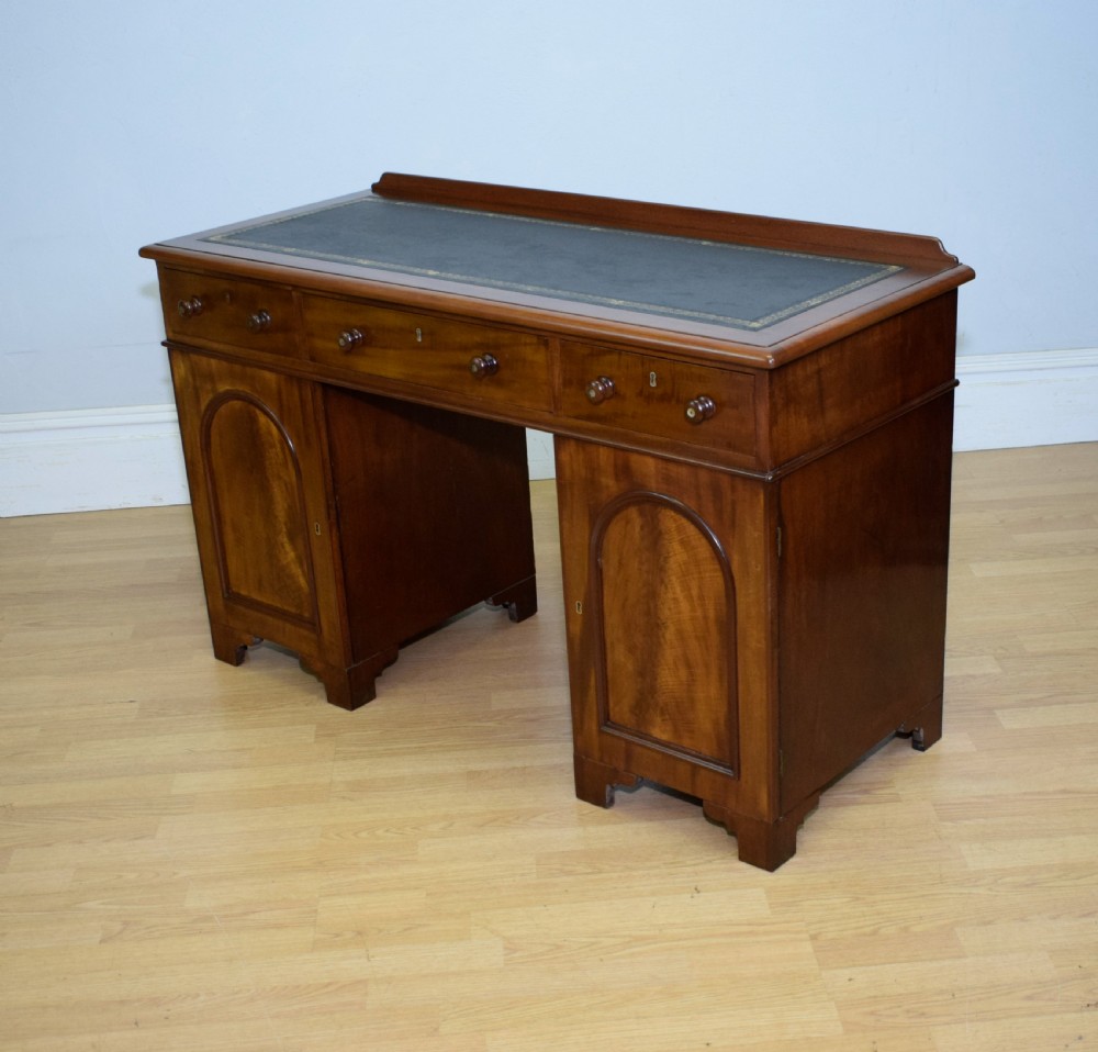 A Victorian mahogany pedestal desk featuring a dark green leather inset writing surface, three frieze drawers, and arched panel pedestal doors. View XVII