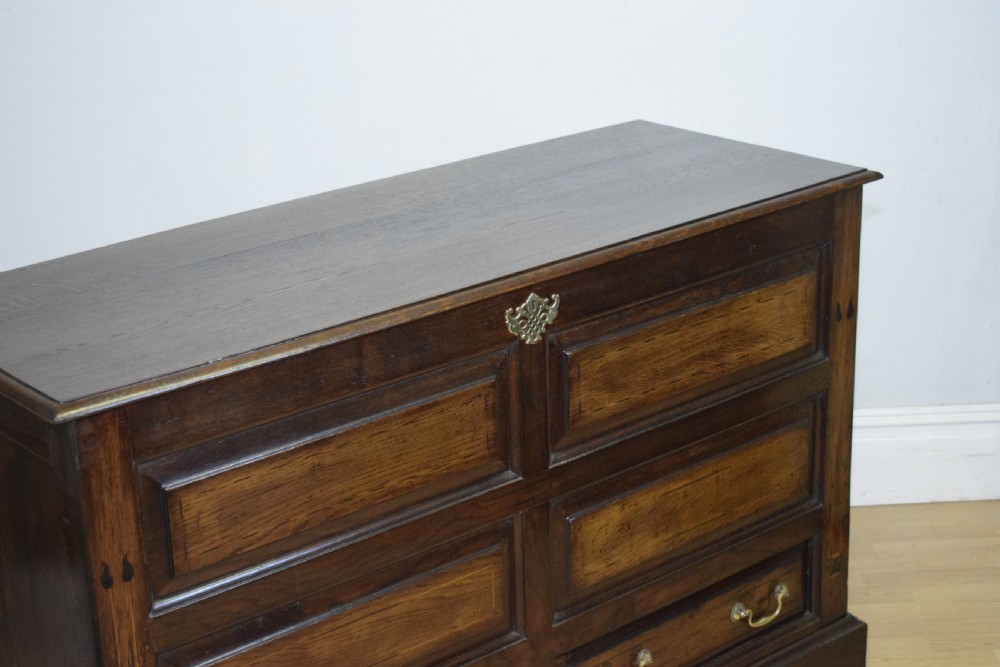 A Georgian dark oak Lancashire mule chest featuring a hinged top lid, panelled front sections with inlays, brass swan-neck handles, and bracket feet. View X