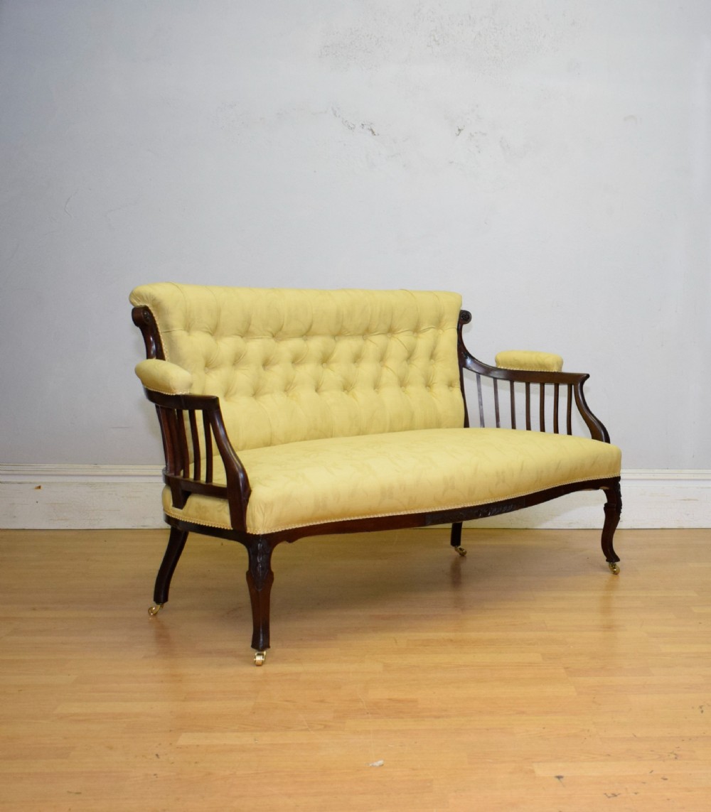 A Victorian mahogany settee featuring yellow tufted upholstery, open spindled arms, carved legs, and brass castors.