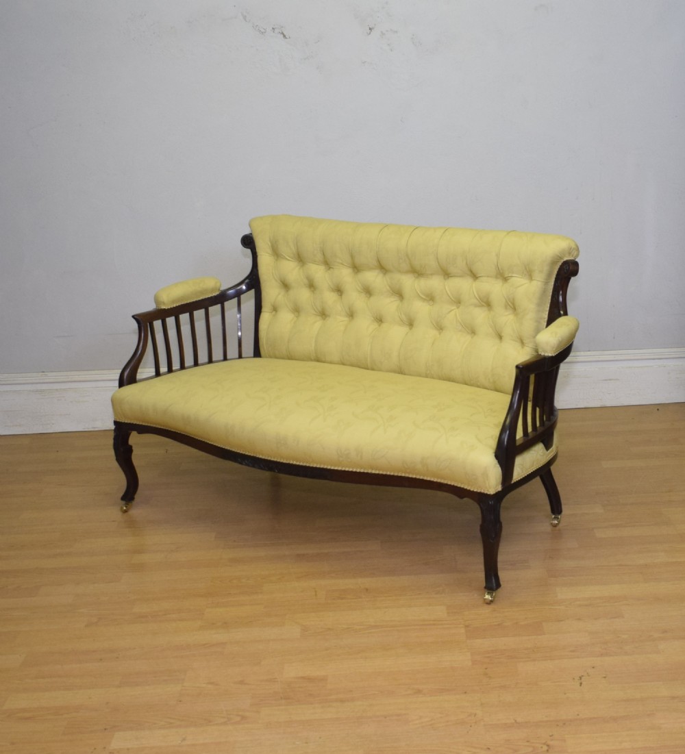 A Victorian mahogany settee featuring yellow tufted upholstery, open spindled arms, carved legs, and brass castors. View VI