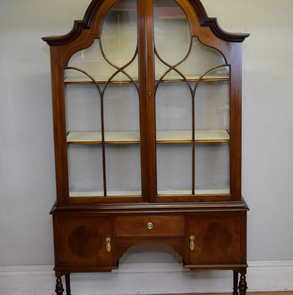 An early 20th-century mahogany display cabinet featuring double astragal glazed doors, an arched pediment top, lower cupboard section, and turned legs with stretchers. View VIII