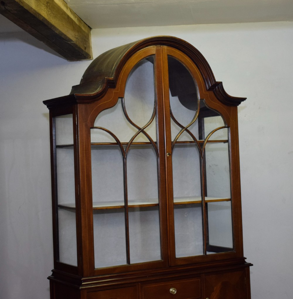 An early 20th-century mahogany display cabinet featuring double astragal glazed doors, an arched pediment top, lower cupboard section, and turned legs with stretchers. View XV