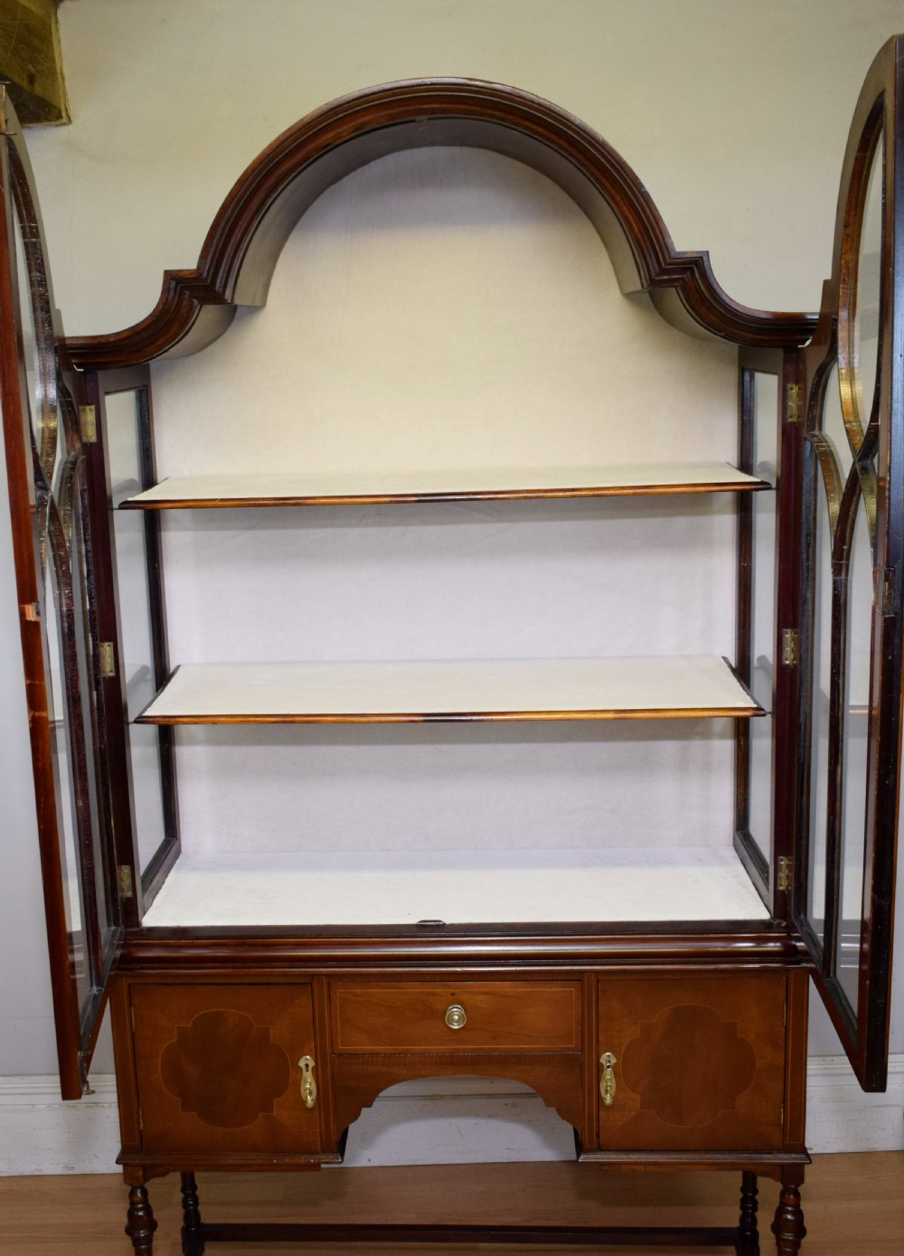 An early 20th-century mahogany display cabinet featuring double astragal glazed doors, an arched pediment top, lower cupboard section, and turned legs with stretchers. View XIX