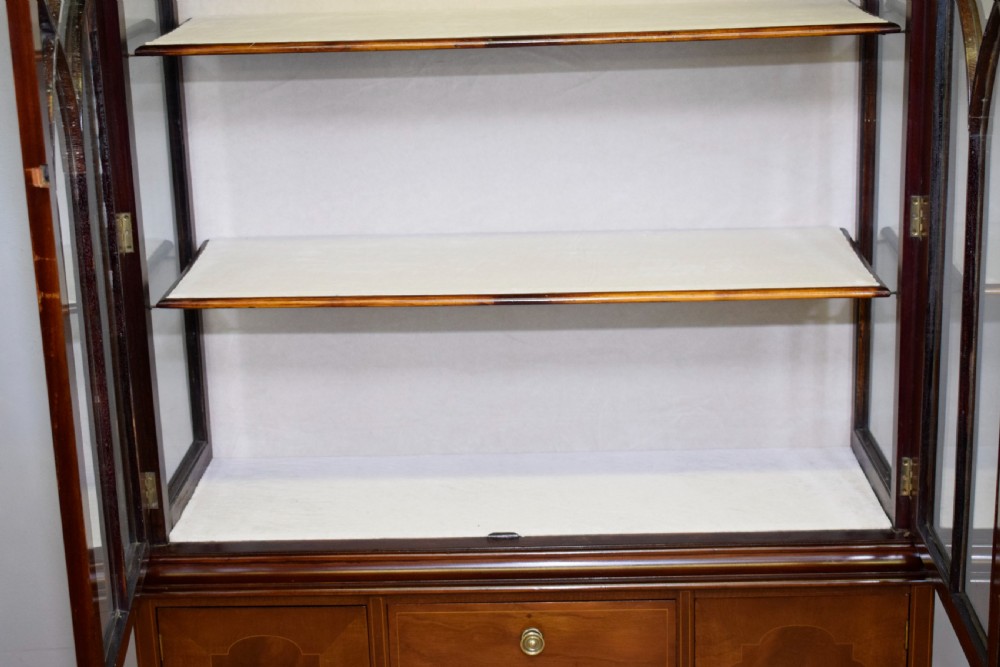 An early 20th-century mahogany display cabinet featuring double astragal glazed doors, an arched pediment top, lower cupboard section, and turned legs with stretchers. View XX