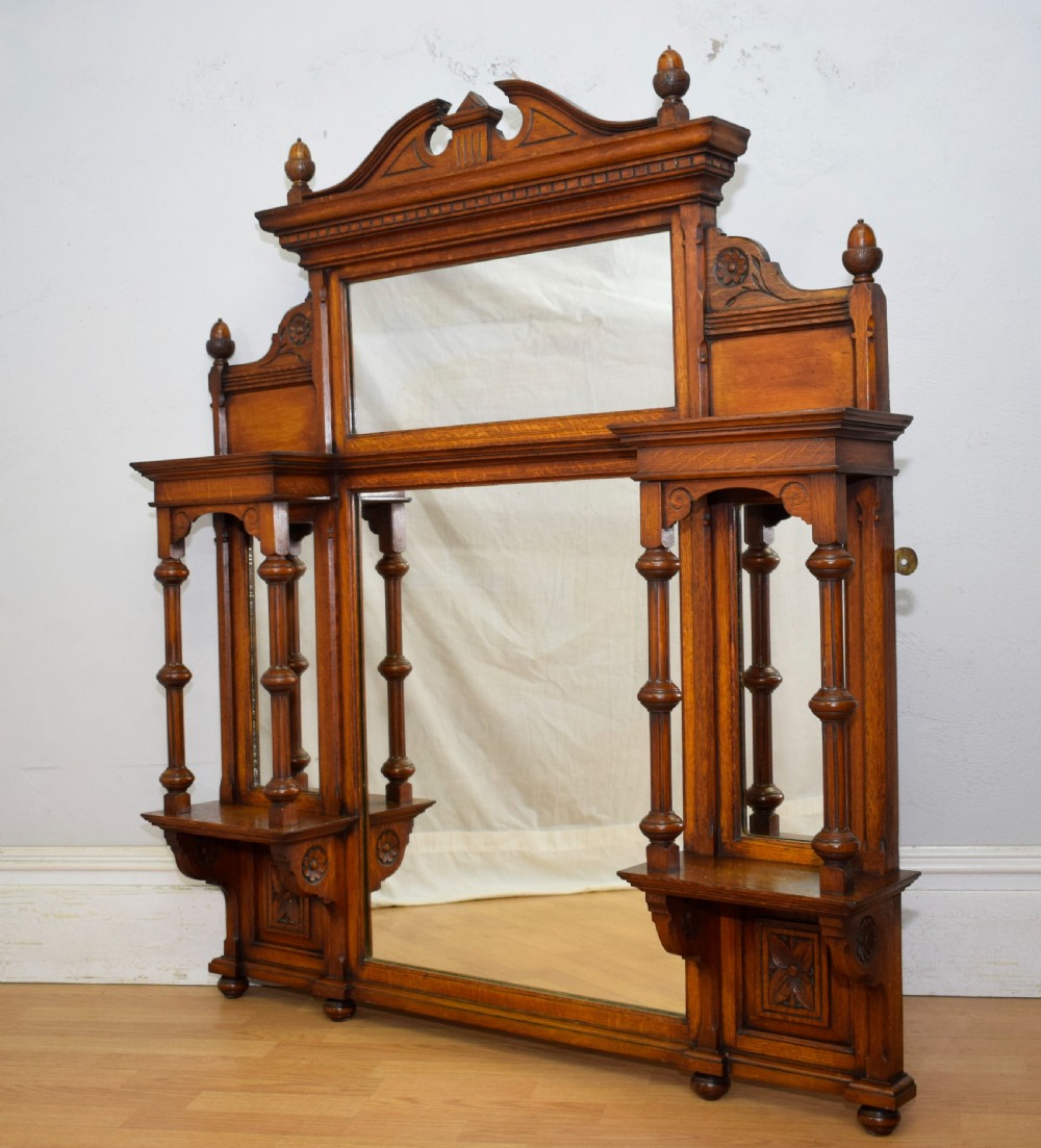 A Victorian carved oak overmantel mirror featuring turned side columns, small side display shelves, an arched pediment, and mirrored glass panels.