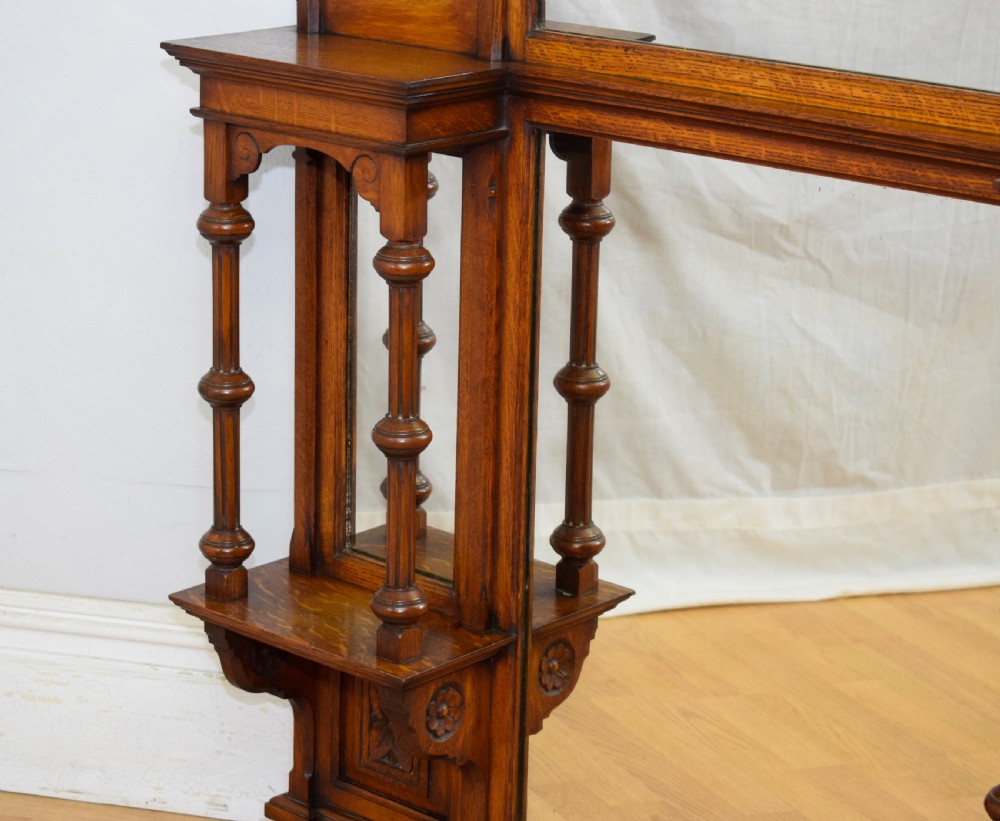 A Victorian carved oak overmantel mirror featuring turned side columns, small side display shelves, an arched pediment, and mirrored glass panels. View X