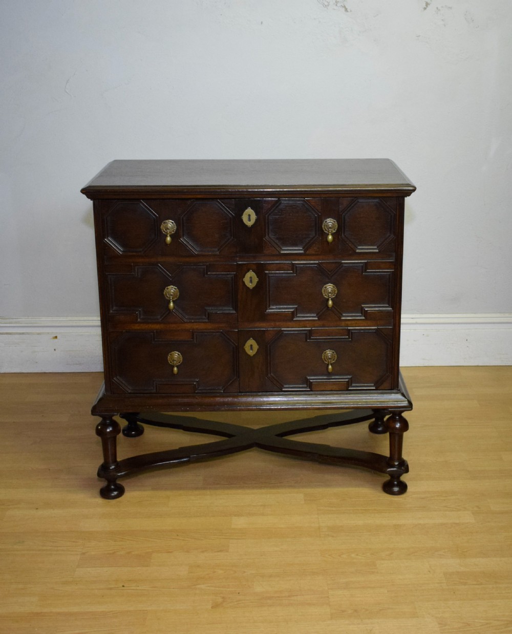 A dark oak Jacobean style cabinet on stand featuring geometric moulded drawer fronts, brass drop handles, turned legs, and an X-shaped stretcher.