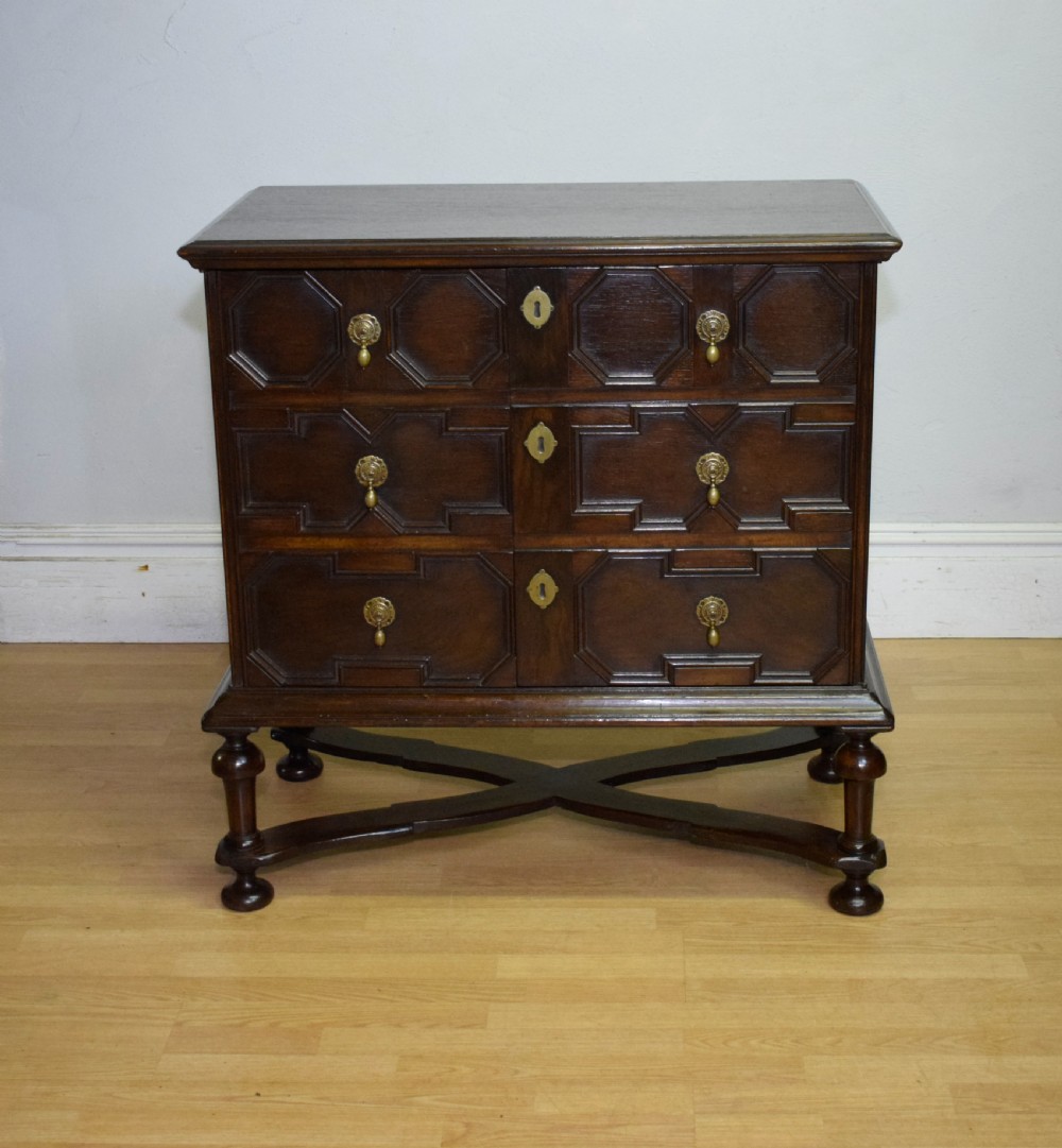 A dark oak Jacobean style cabinet on stand featuring geometric moulded drawer fronts, brass drop handles, turned legs, and an X-shaped stretcher. View III