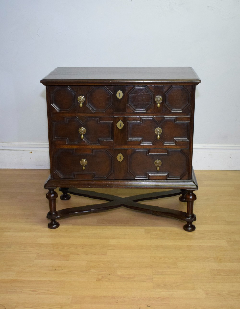 A dark oak Jacobean style cabinet on stand featuring geometric moulded drawer fronts, brass drop handles, turned legs, and an X-shaped stretcher. View V