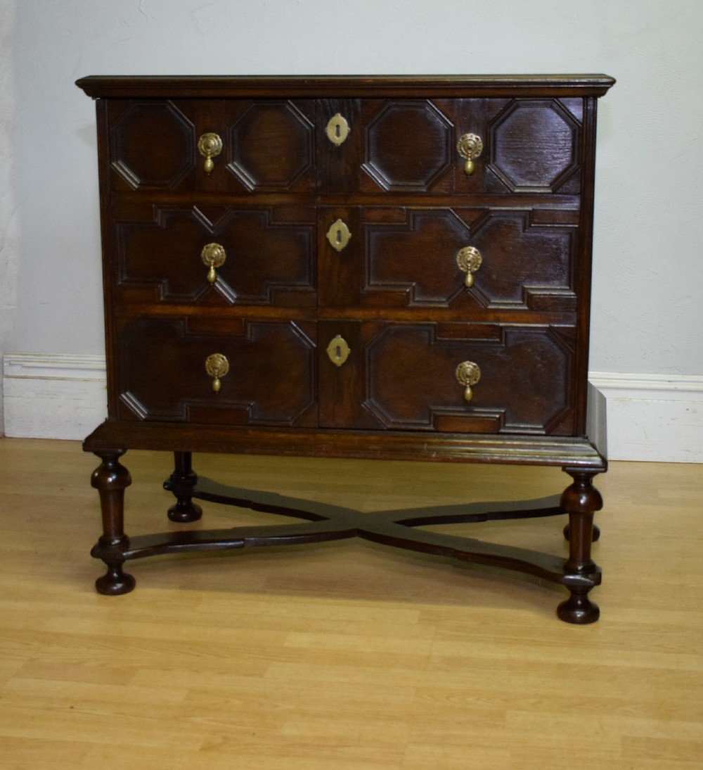 A dark oak Jacobean style cabinet on stand featuring geometric moulded drawer fronts, brass drop handles, turned legs, and an X-shaped stretcher. View VI