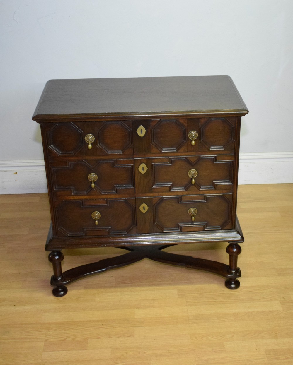A dark oak Jacobean style cabinet on stand featuring geometric moulded drawer fronts, brass drop handles, turned legs, and an X-shaped stretcher. View VII