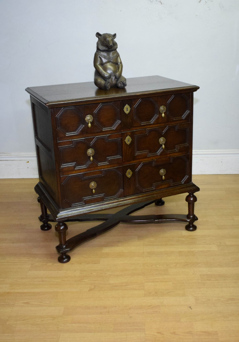 A dark oak Jacobean style cabinet on stand featuring geometric moulded drawer fronts, brass drop handles, turned legs, and an X-shaped stretcher. View X