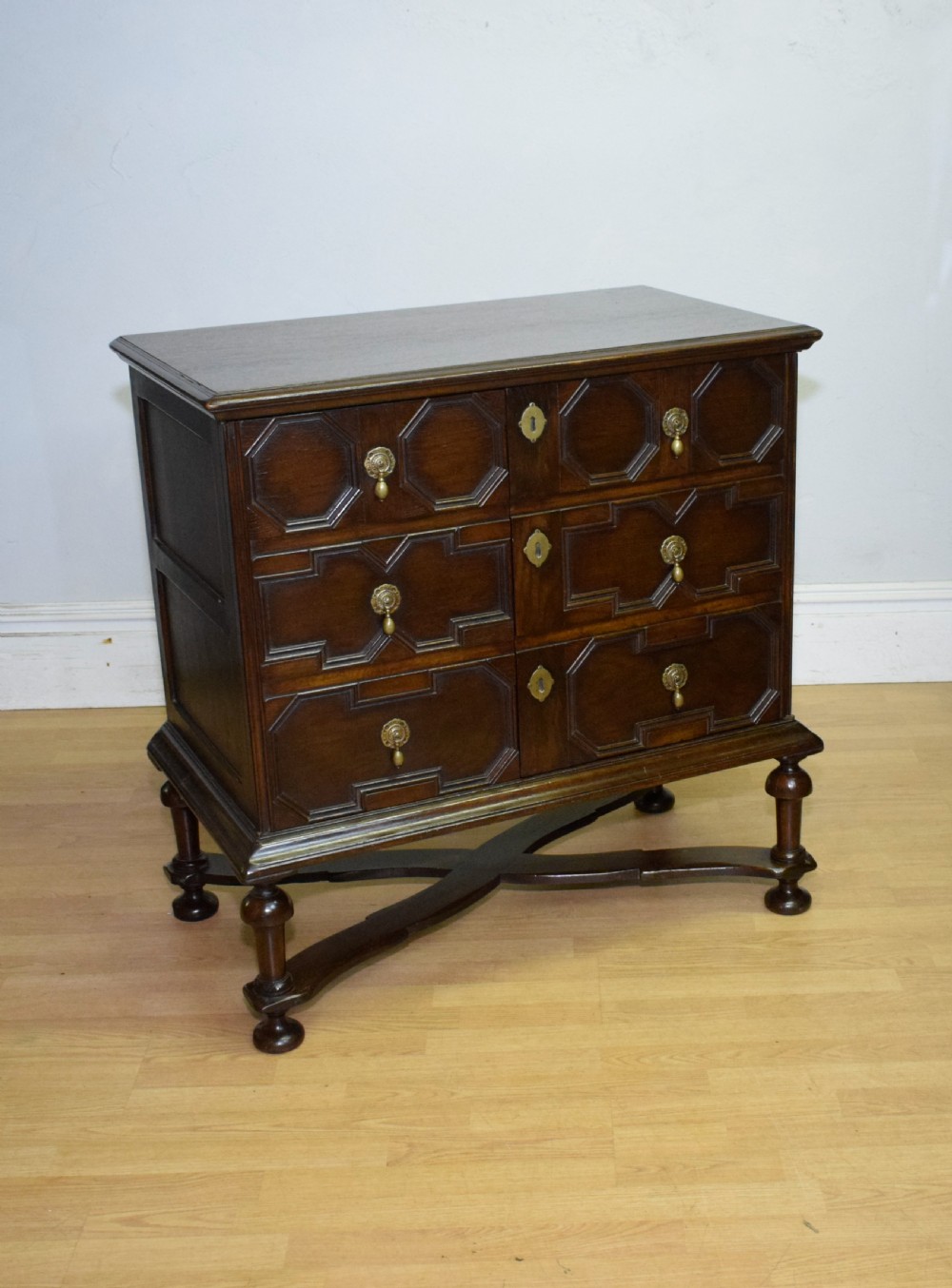 A dark oak Jacobean style cabinet on stand featuring geometric moulded drawer fronts, brass drop handles, turned legs, and an X-shaped stretcher. View XII