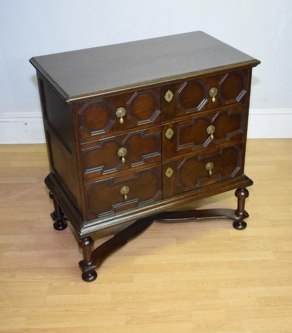 A dark oak Jacobean style cabinet on stand featuring geometric moulded drawer fronts, brass drop handles, turned legs, and an X-shaped stretcher. View XIII
