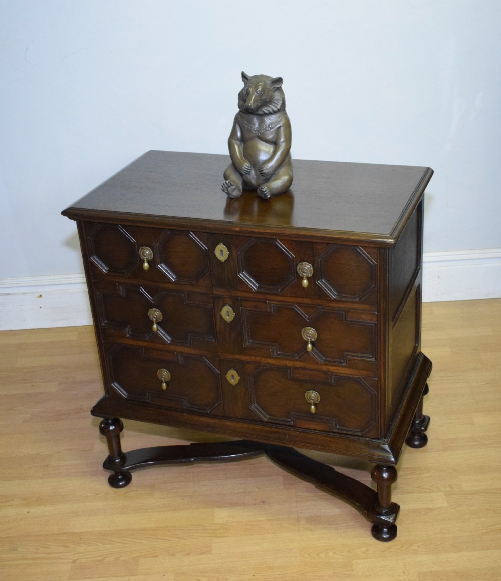 A dark oak Jacobean style cabinet on stand featuring geometric moulded drawer fronts, brass drop handles, turned legs, and an X-shaped stretcher. View XV