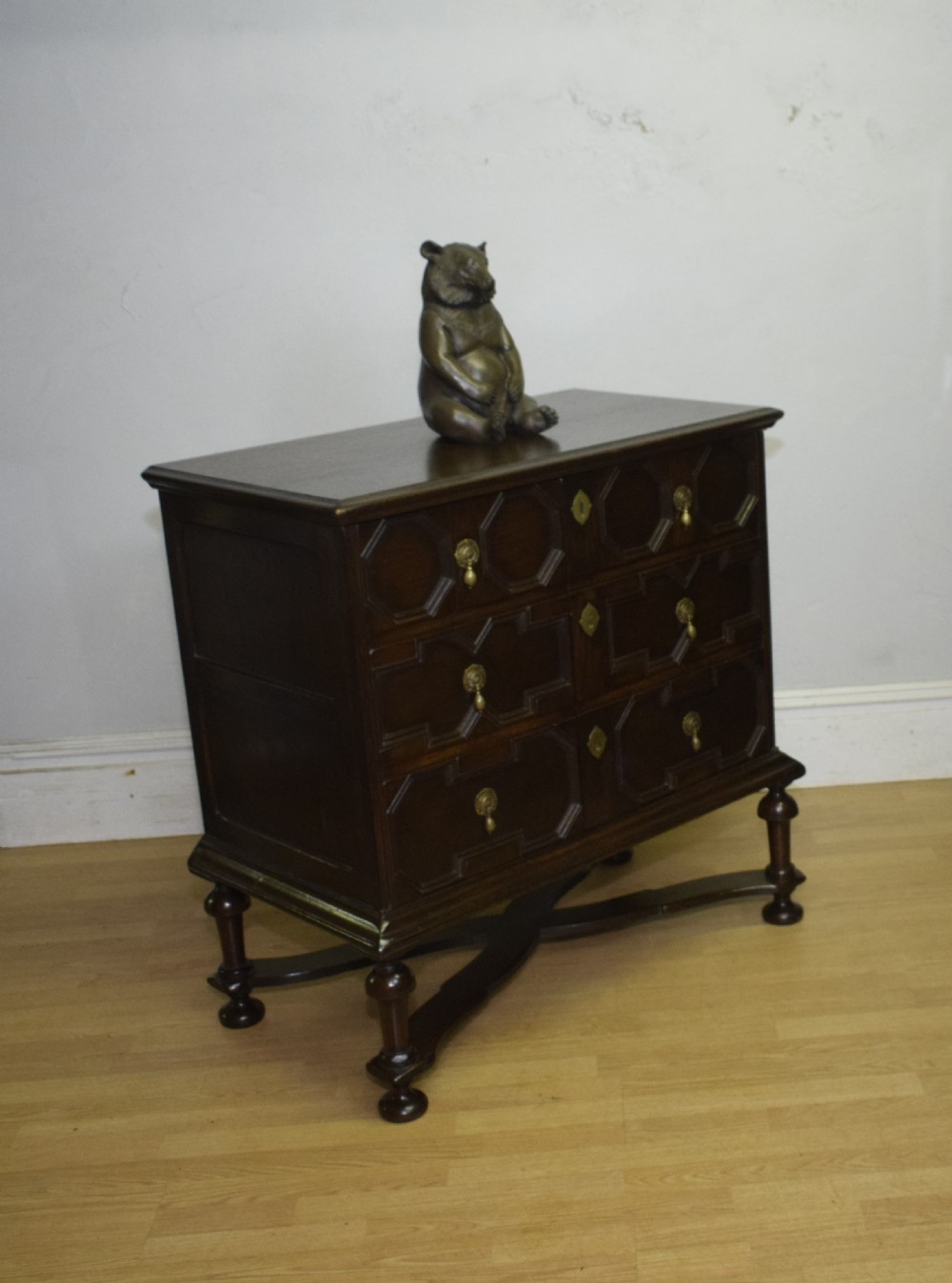 A dark oak Jacobean style cabinet on stand featuring geometric moulded drawer fronts, brass drop handles, turned legs, and an X-shaped stretcher. View XVIII