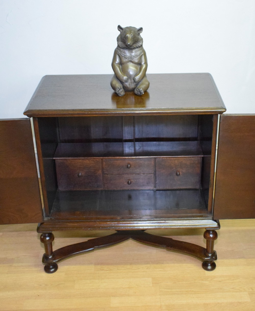 A dark oak Jacobean style cabinet on stand featuring geometric moulded drawer fronts, brass drop handles, turned legs, and an X-shaped stretcher. View XXI