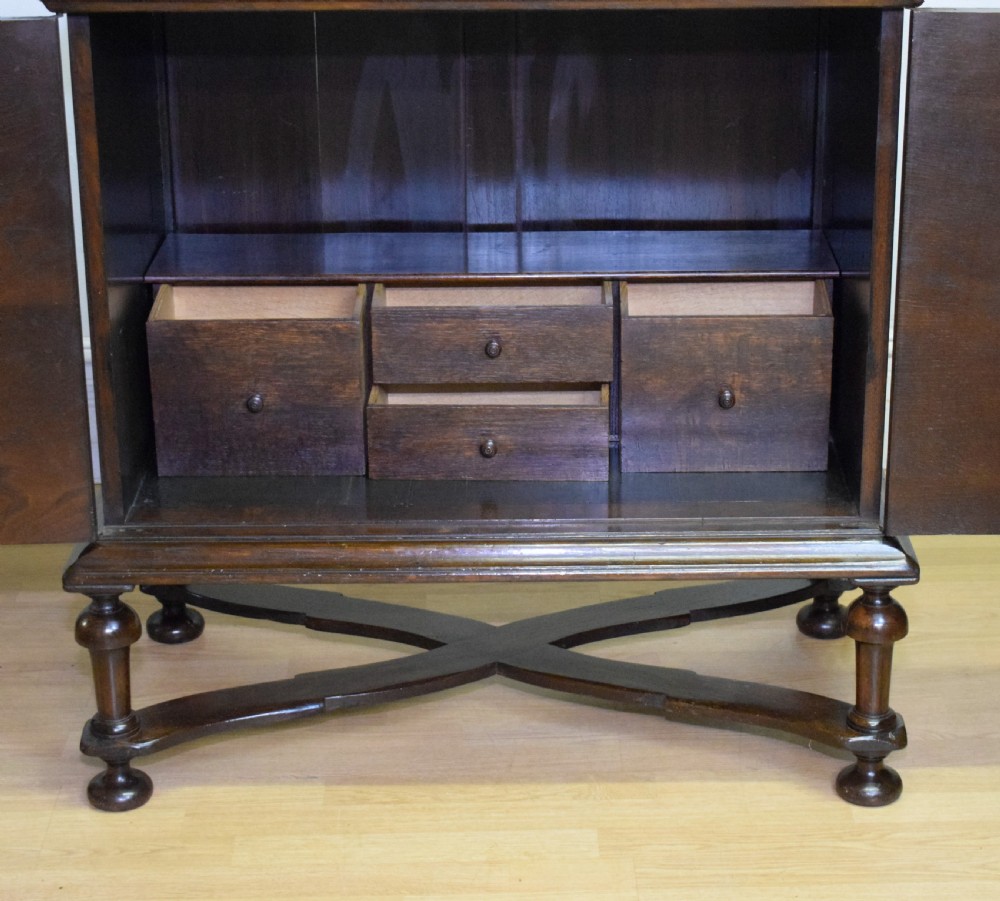 A dark oak Jacobean style cabinet on stand featuring geometric moulded drawer fronts, brass drop handles, turned legs, and an X-shaped stretcher. View XXII