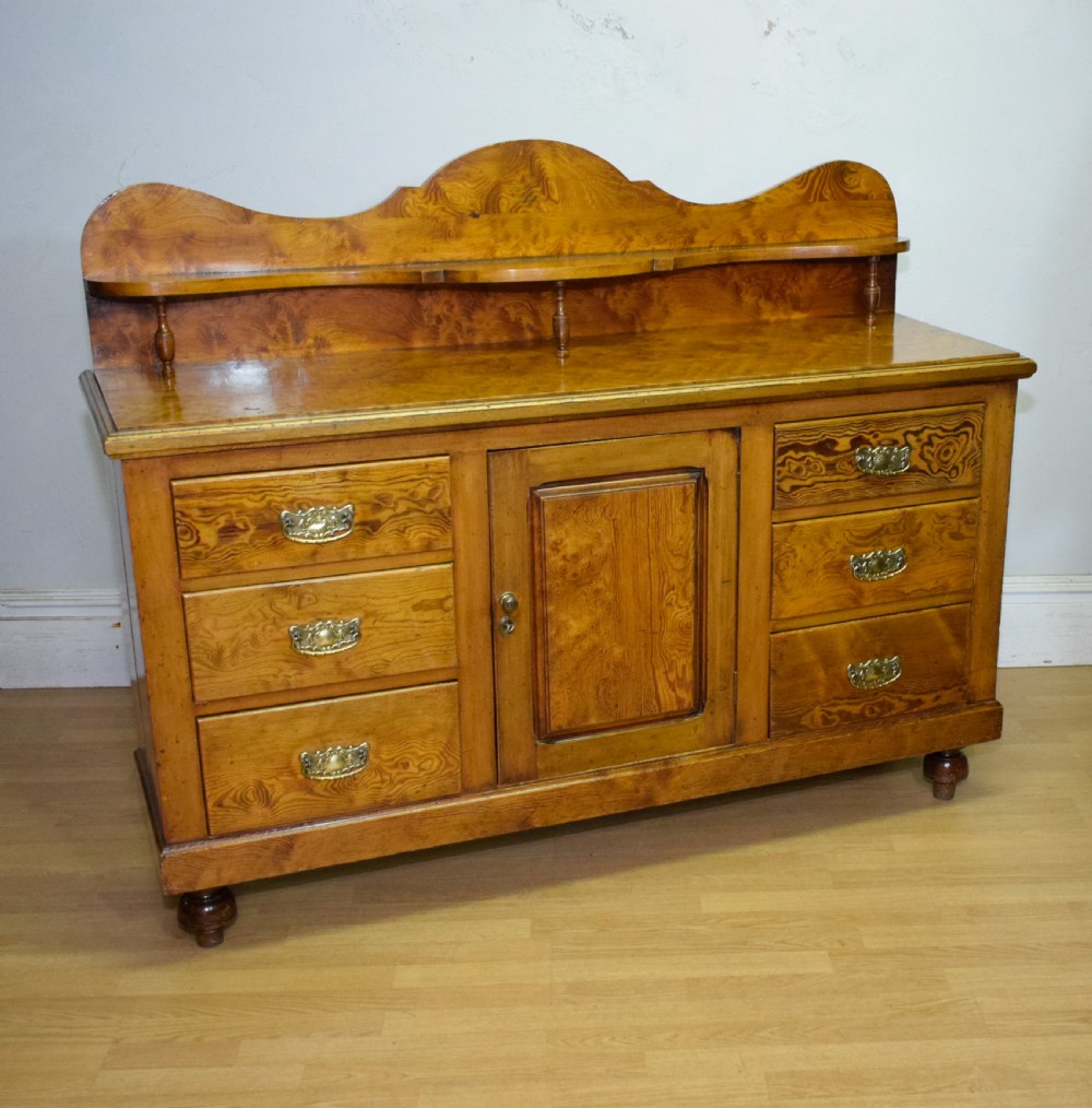 A Victorian golden pine dresser base featuring six drawers, a central panelled cupboard door, brass handles, and a shaped gallery back with a small shelf.