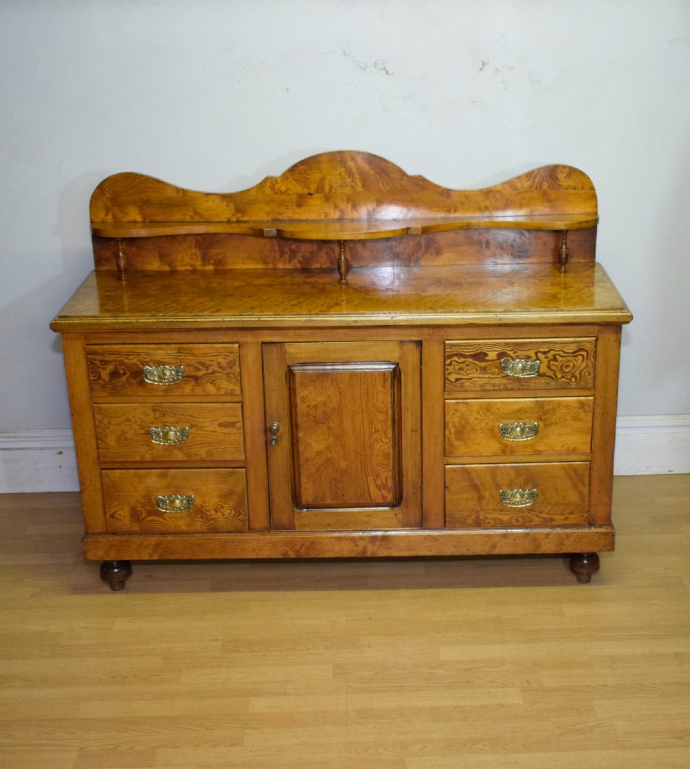 A Victorian golden pine dresser base featuring six drawers, a central panelled cupboard door, brass handles, and a shaped gallery back with a small shelf. View II