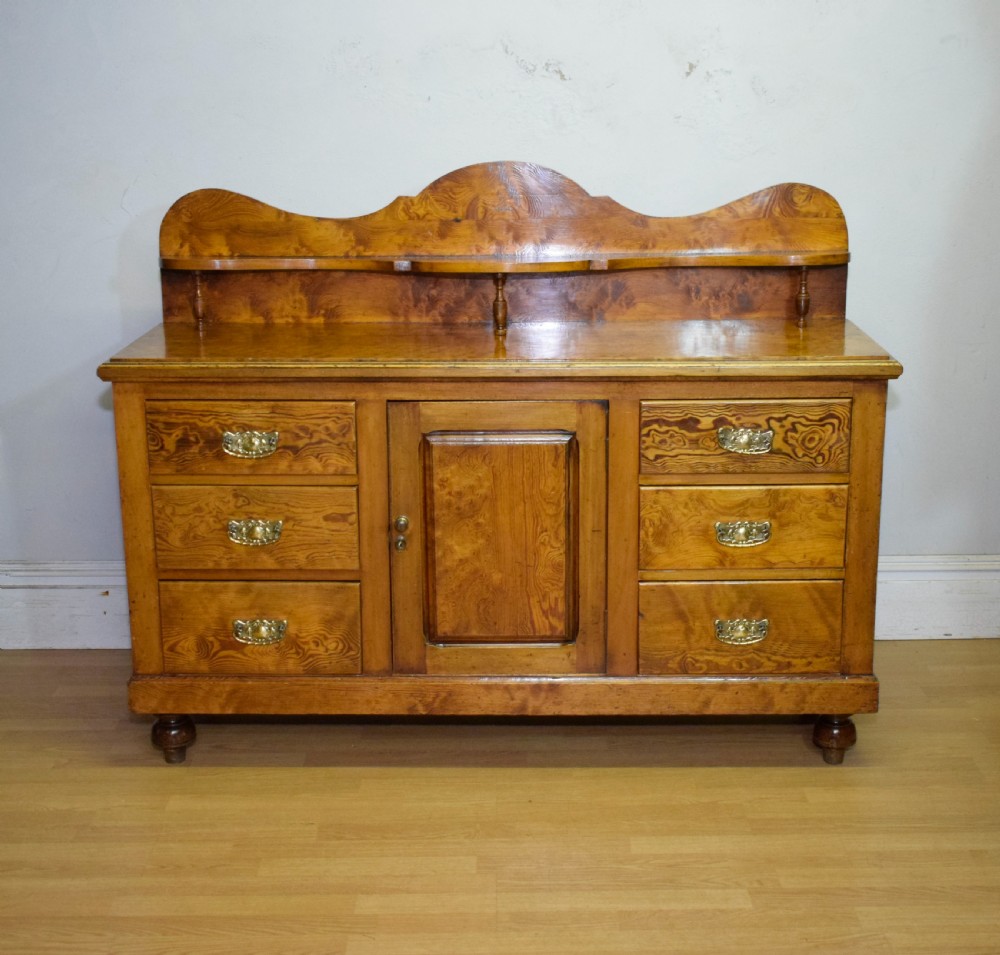 A Victorian golden pine dresser base featuring six drawers, a central panelled cupboard door, brass handles, and a shaped gallery back with a small shelf. View III