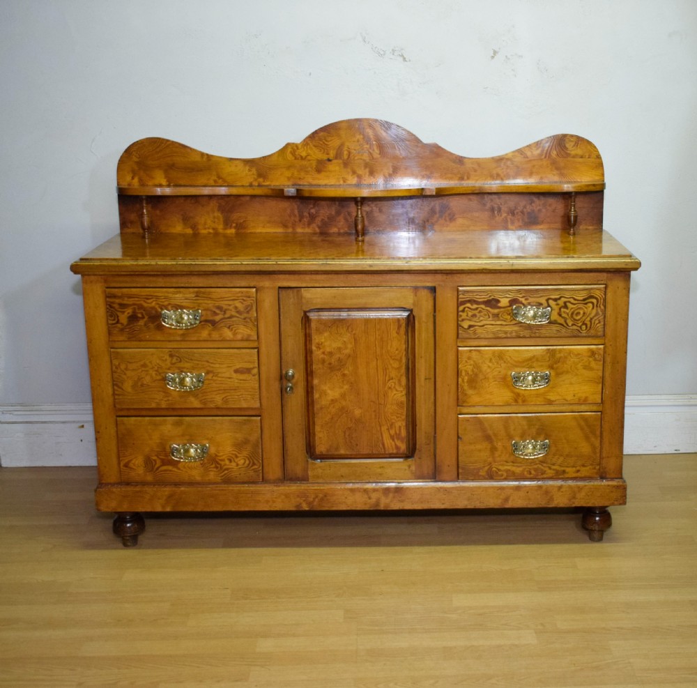 A Victorian golden pine dresser base featuring six drawers, a central panelled cupboard door, brass handles, and a shaped gallery back with a small shelf. View V
