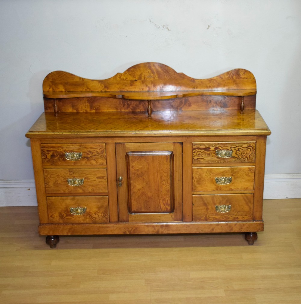 A Victorian golden pine dresser base featuring six drawers, a central panelled cupboard door, brass handles, and a shaped gallery back with a small shelf. View VI