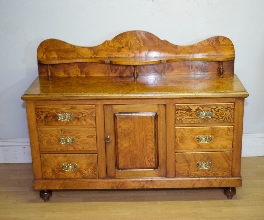 A Victorian golden pine dresser base featuring six drawers, a central panelled cupboard door, brass handles, and a shaped gallery back with a small shelf. View VIII