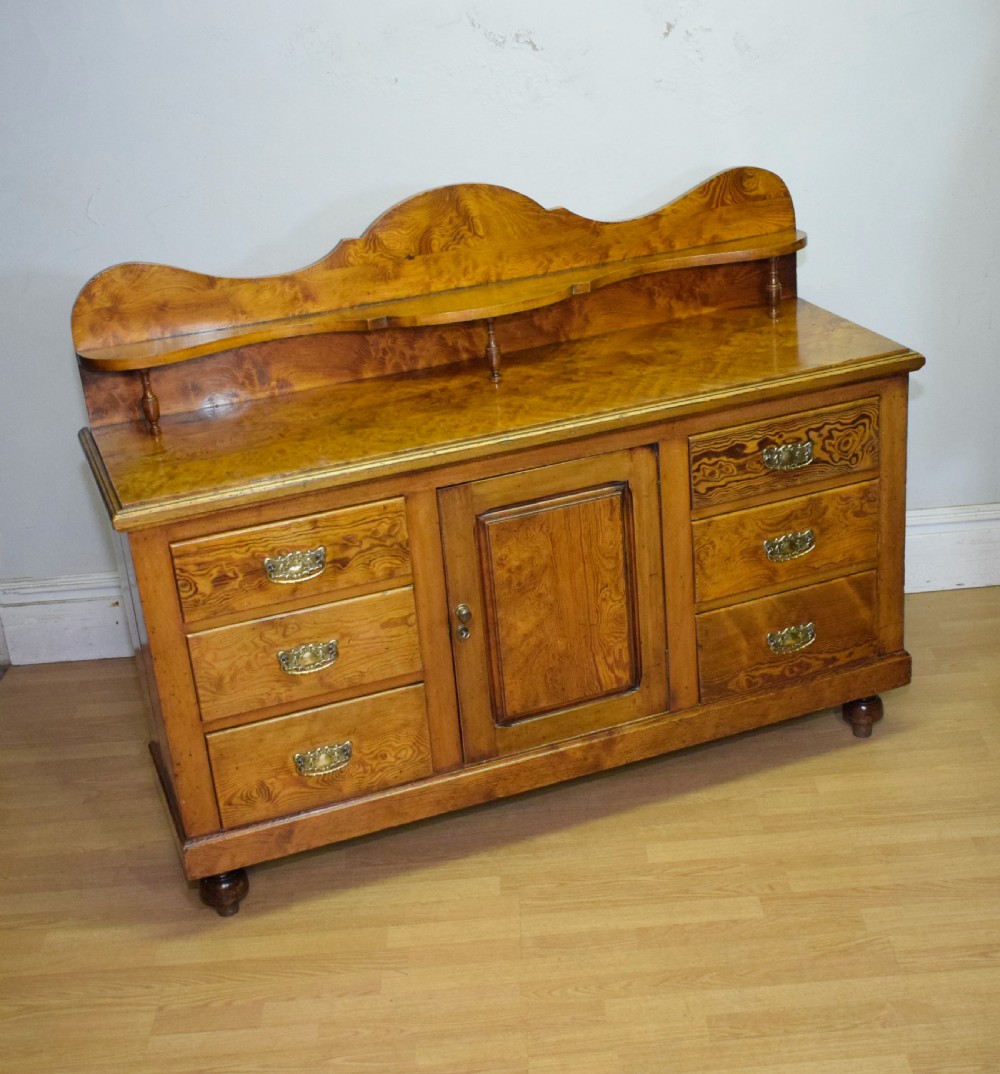 A Victorian golden pine dresser base featuring six drawers, a central panelled cupboard door, brass handles, and a shaped gallery back with a small shelf. View X