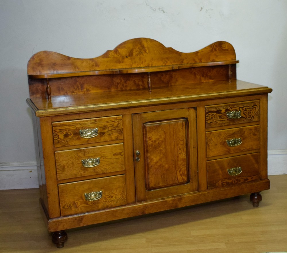 A Victorian golden pine dresser base featuring six drawers, a central panelled cupboard door, brass handles, and a shaped gallery back with a small shelf. View XII