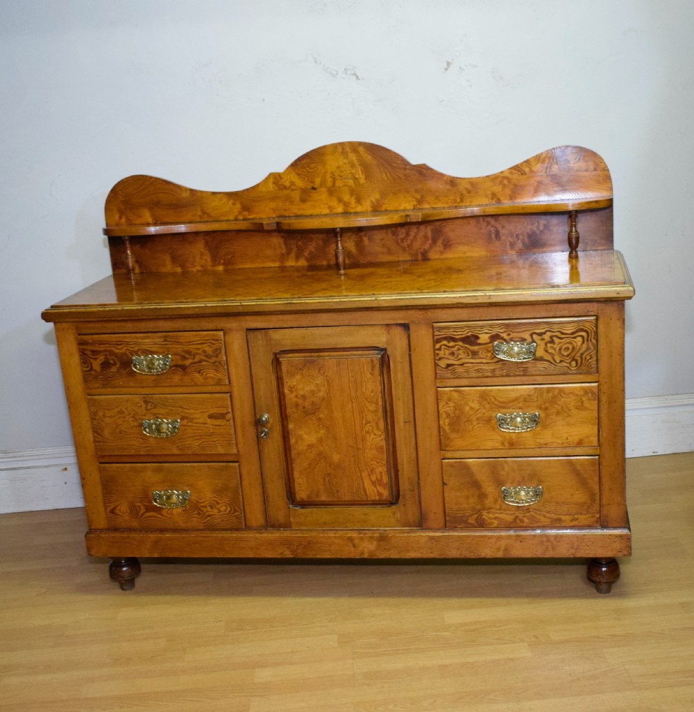 A Victorian golden pine dresser base featuring six drawers, a central panelled cupboard door, brass handles, and a shaped gallery back with a small shelf. View XIV