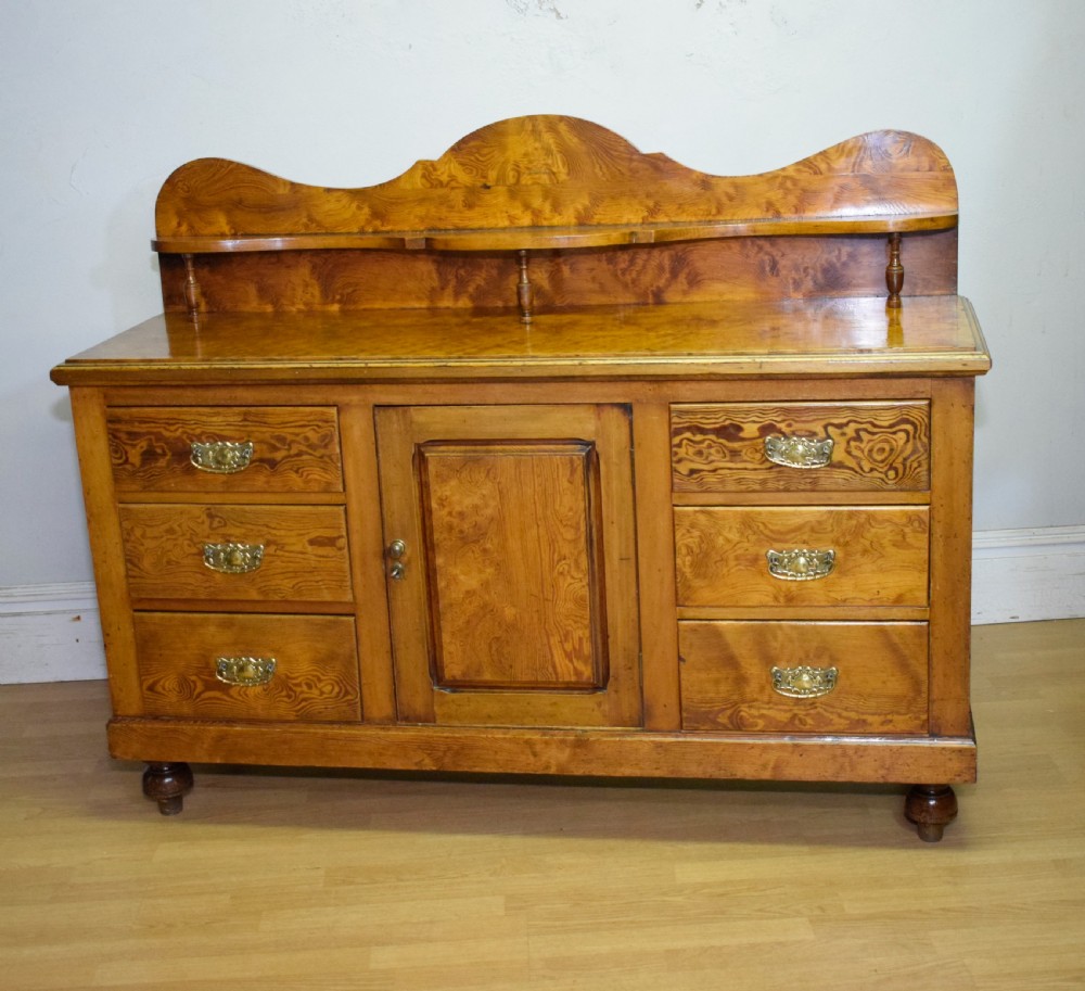 A Victorian golden pine dresser base featuring six drawers, a central panelled cupboard door, brass handles, and a shaped gallery back with a small shelf. View XV