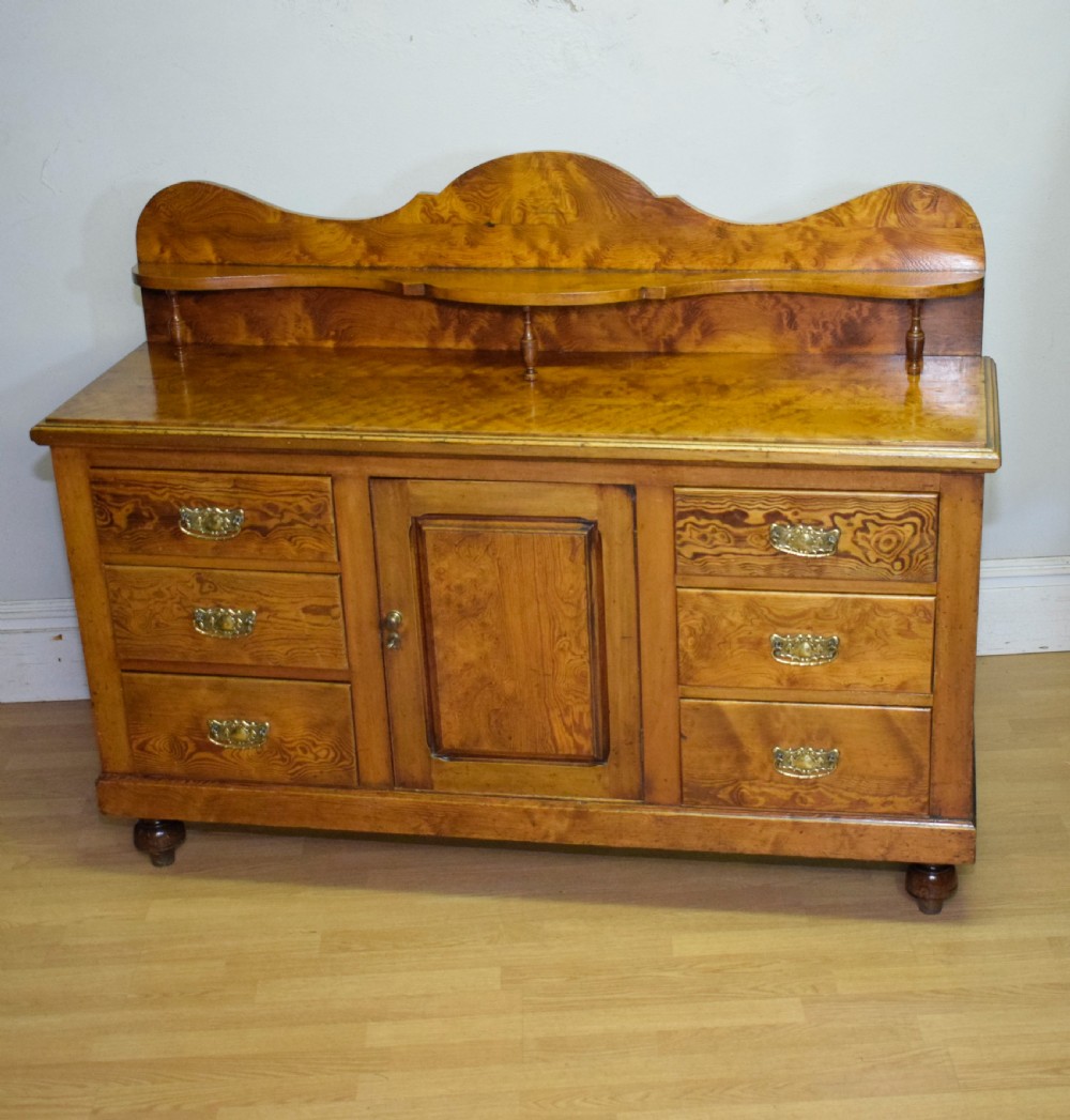 A Victorian golden pine dresser base featuring six drawers, a central panelled cupboard door, brass handles, and a shaped gallery back with a small shelf. View XVI