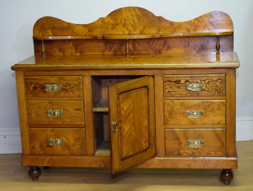 A Victorian golden pine dresser base featuring six drawers, a central panelled cupboard door, brass handles, and a shaped gallery back with a small shelf. View XVII