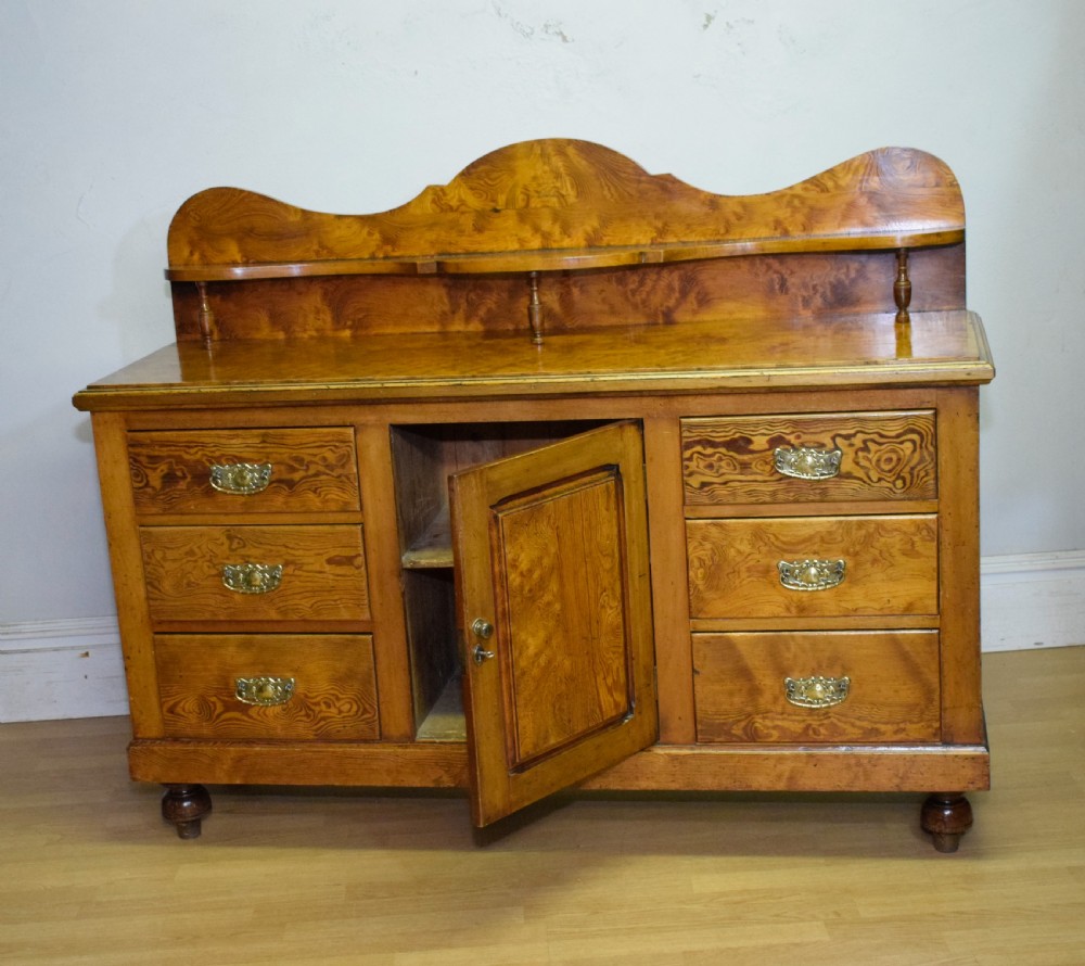A Victorian golden pine dresser base featuring six drawers, a central panelled cupboard door, brass handles, and a shaped gallery back with a small shelf. View XVIII
