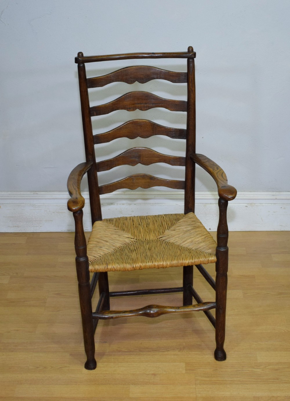A 19th-century dark oak ladder-back armchair featuring five shaped horizontal back slats, turned legs, curved armrests, and a woven rush seat.