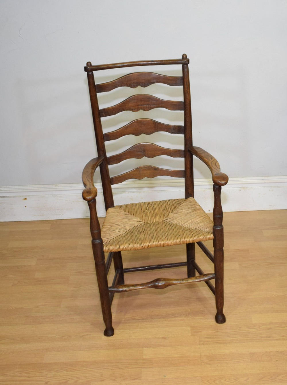 A 19th-century dark oak ladder-back armchair featuring five shaped horizontal back slats, turned legs, curved armrests, and a woven rush seat. View III