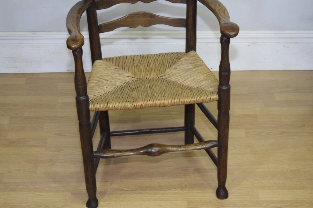 A 19th-century dark oak ladder-back armchair featuring five shaped horizontal back slats, turned legs, curved armrests, and a woven rush seat. View V